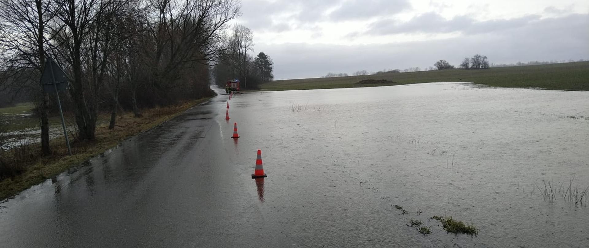 Na zdjęciu duże rozlewisko z prawej strony. Woda przelewa się przez drogę. W oddali samochód strażacki.