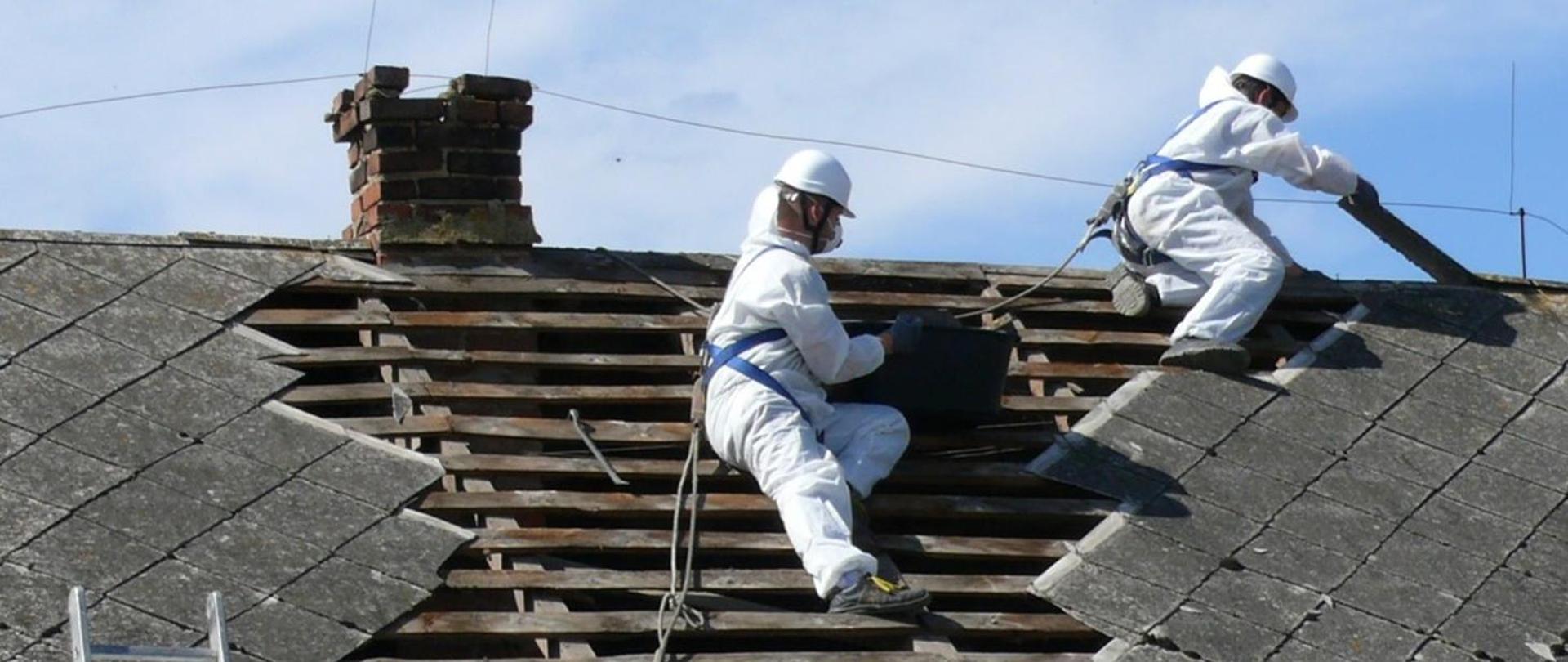 Męźczyźni w kombinezonach, kaskach, butach roboczych i maseczkach, zabezpieczeni linami usuwają zniszczone płyty azbestowe z dachu