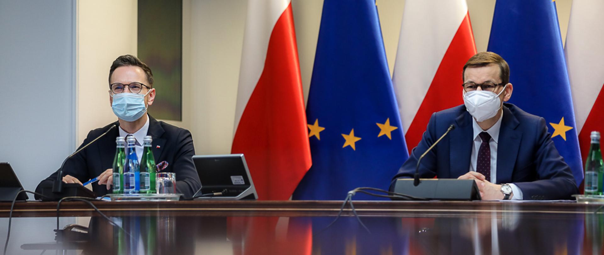Premier Mateusz Morawiecki i wiceminister Waldemar Buda siedzą przy stole. W tle flagi Polski i UE.