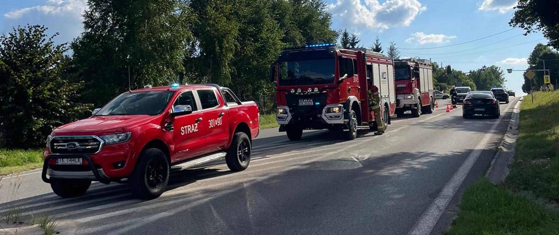 Zdjęcie przedstawia drogę powiatową gdzie na jednym pasie stoją kolejno trzy wozy strażackie. W głębi widać pachołki ostrzegawcze wytyczające teren działań. Drugim pasem przejeżdżają przypadkowe auta.