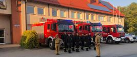 Strażacy stoją w szeregu przed komendą Państwowej Straży Pożarnej, za nimi widoczne samochody gaśnicze i operacyjny, dwóch strażaków w mundurach koloru musztardowego salutuje