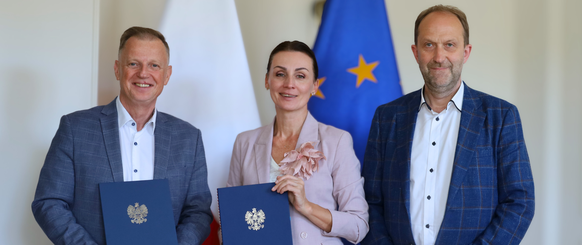 trzy osoby trzymają w rękach podpisane umowy pozują do zdjęcia na tle flag