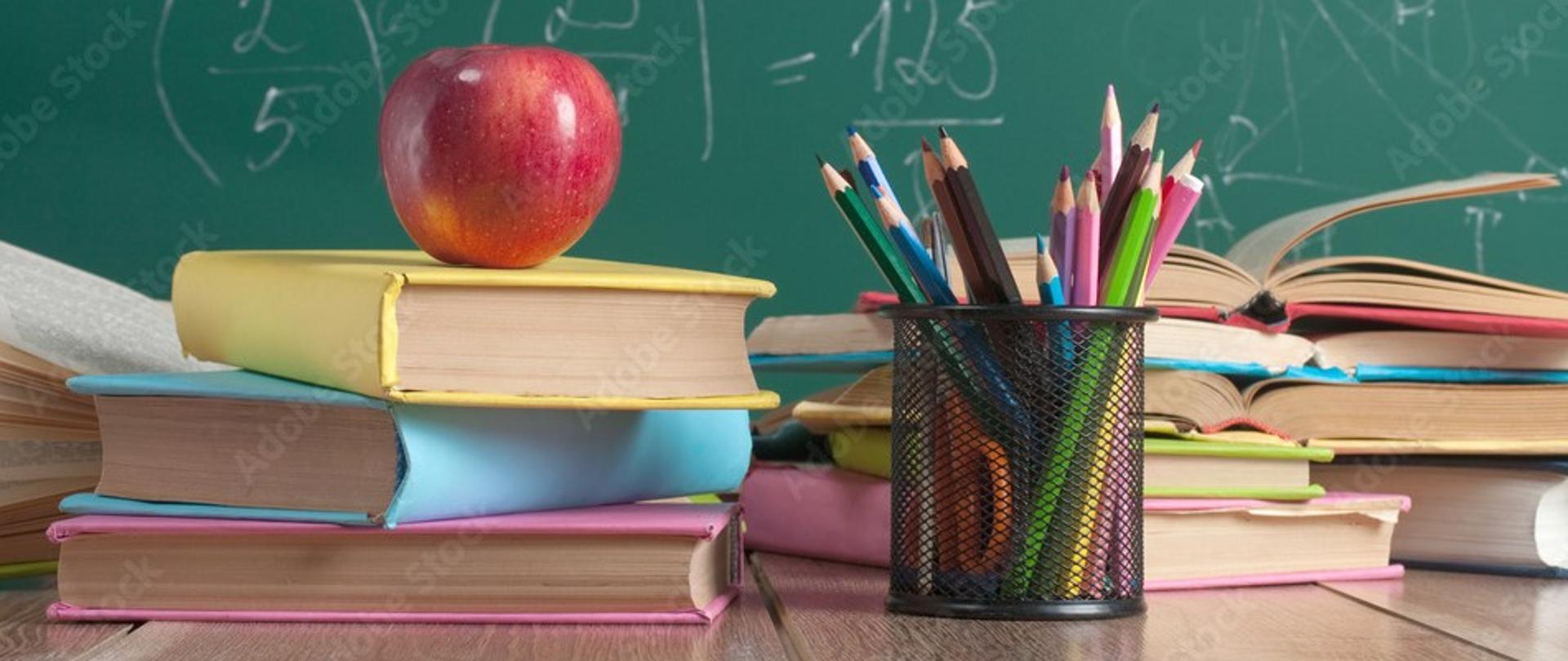 Na tle zielonej szkolnej tablicy na biurku stos książek , zbór kolorowych kredek w pojemniku i czerwone jabłko na szczycie książek