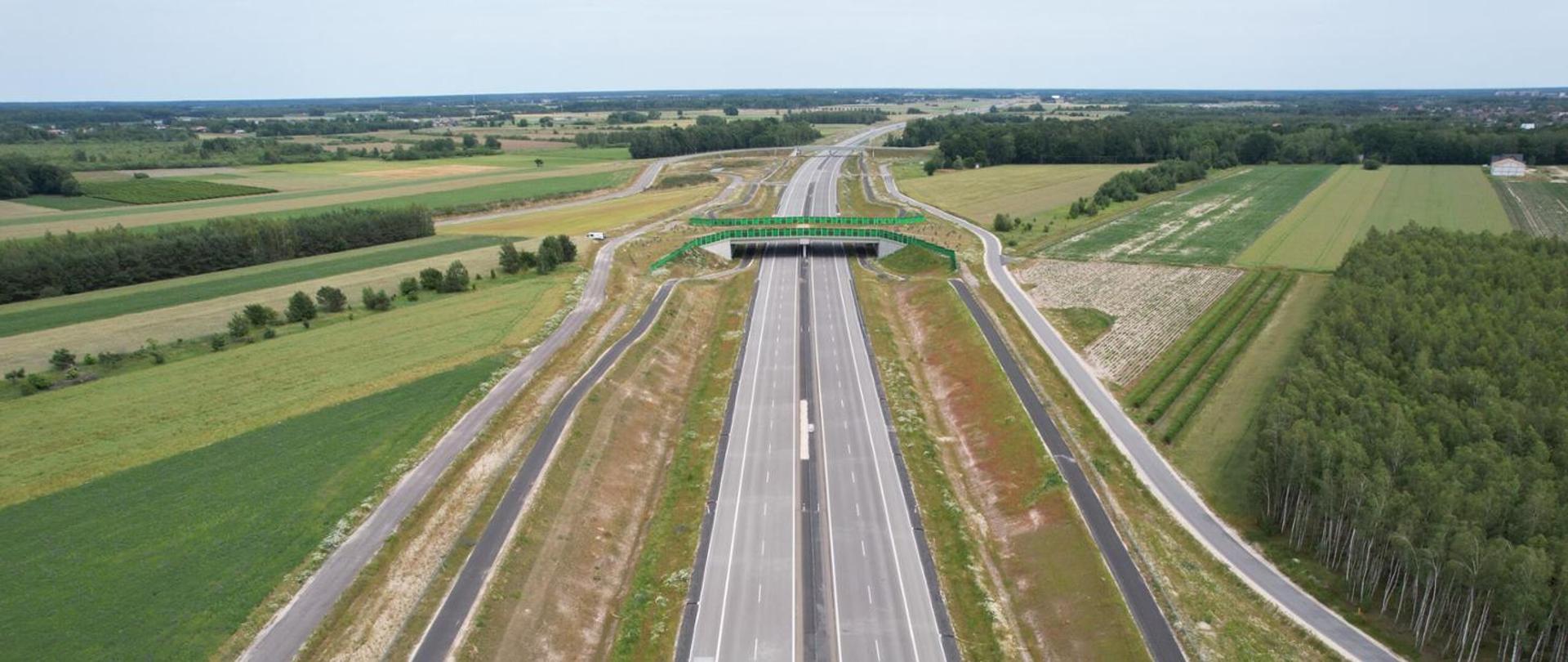 Widok z lotu ptaka na nową dwujezdniową drogę przebiegającą przez tereny rolnicze; nad jezdniami zielone przejście dla zwierząt, po obu stronach drogi serwisowe, w tle pola i las.
