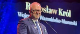 Mężczyzna w garniturze stoi za mównicą na tle ścianki granatowej 