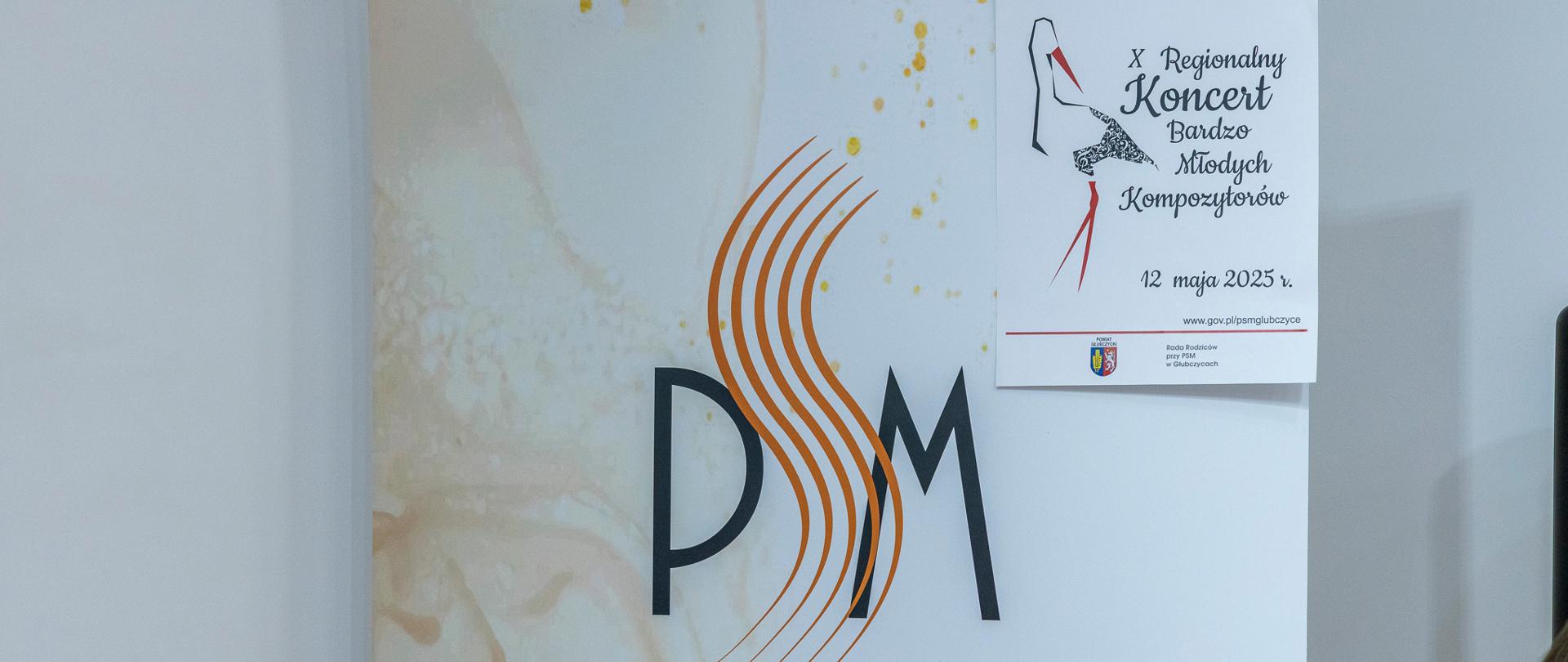 Baner szkolny stojący na scenie, na banerze logo szkoły i przyczepiony w jego górnym prawym rogu plakatem informującym o dziesiątym koncercie bardzo młodych kompozytorów.