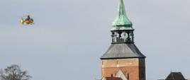 Zdjęcie przedstawia lądujący przy kościele mariackim w Białogardzie śmigłowiec Lotniczego Pogotowia Ratowniczego. Żółty śmigłowiec unosi się nad dachami budynków mieszkalnych