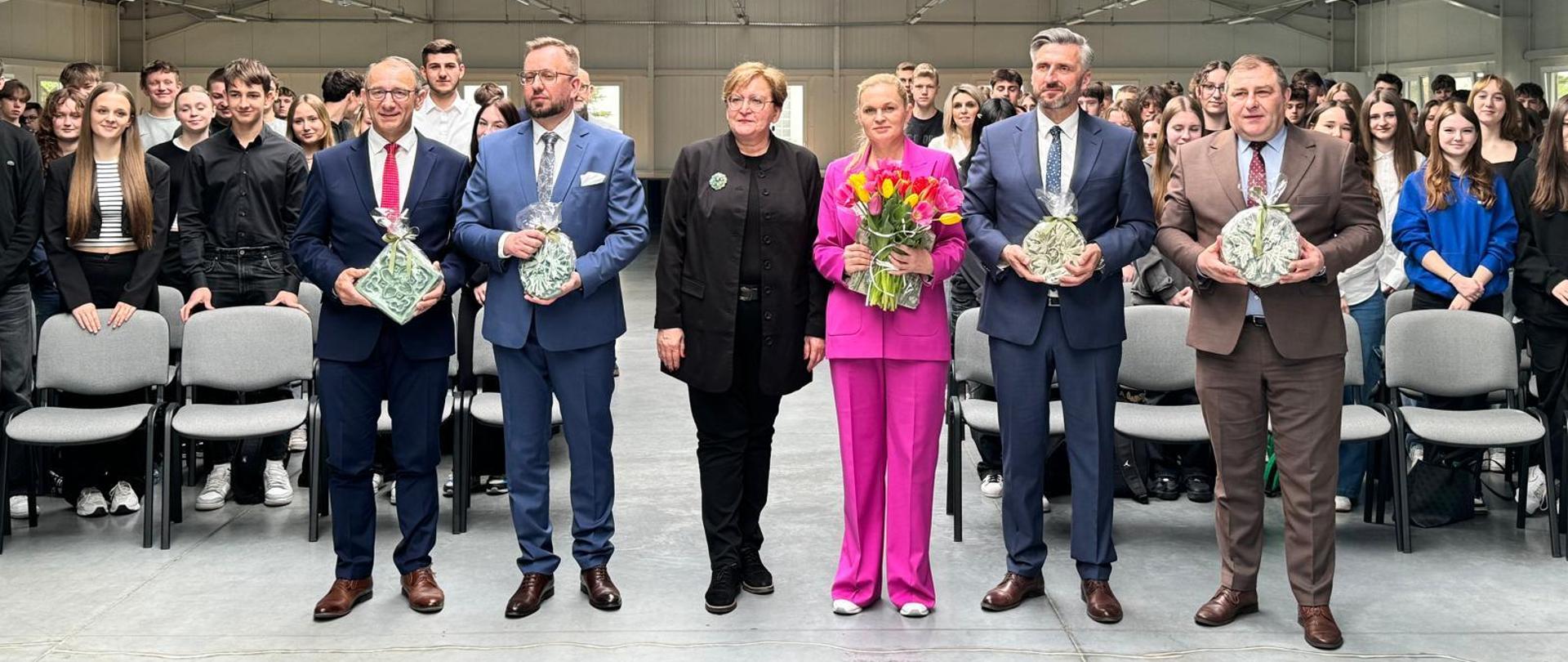 Minister Barbara Nowacka wraz z innymi gośćmi pozuje do zdjęcia na stojąco. w tle uczniowie i nauczyciele siedzący na krzesłach. Minister trzyma w ręku kolorowe tulipany. Ubrana w różowy garnitur. Wizyta minister Barbary Nowackiej w województwie lubuskim