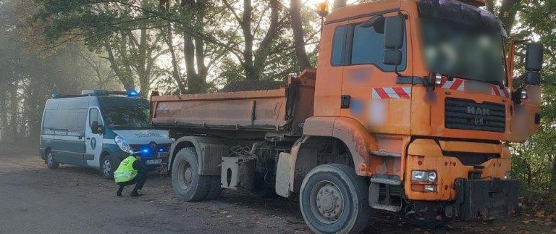 Pomarańczowa wywrotka. Inspektor schylony kontroluje stan ogumienia tylnej osi. Za pojazdem radiowóz ITD.