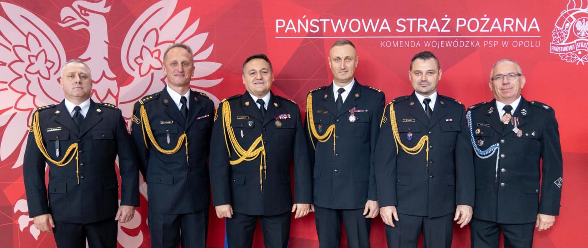 Na zdjęciu znajdują się strażacy ubrani w mundury, w tle znajduje się baner z logo PSP