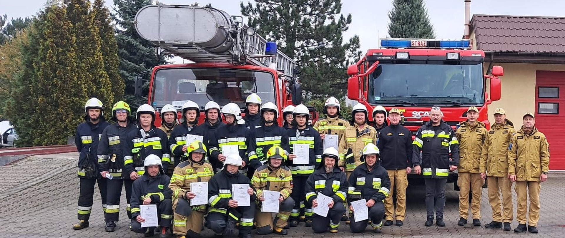 Zdjęcie przedstawia zbiórkę strażaków OSP w umundurowaniu specjalnym wraz z funkcjonariuszami z PSP nadzorującymi szkolenie oraz Prezesem Zarządu Oddziału Powiatowego ZOSP RP. Zbiórka jest zrealizowana na tle samochodów pożarniczych SCD z JRG Zduńska Wola oraz GCBA z OSP Zapolice.