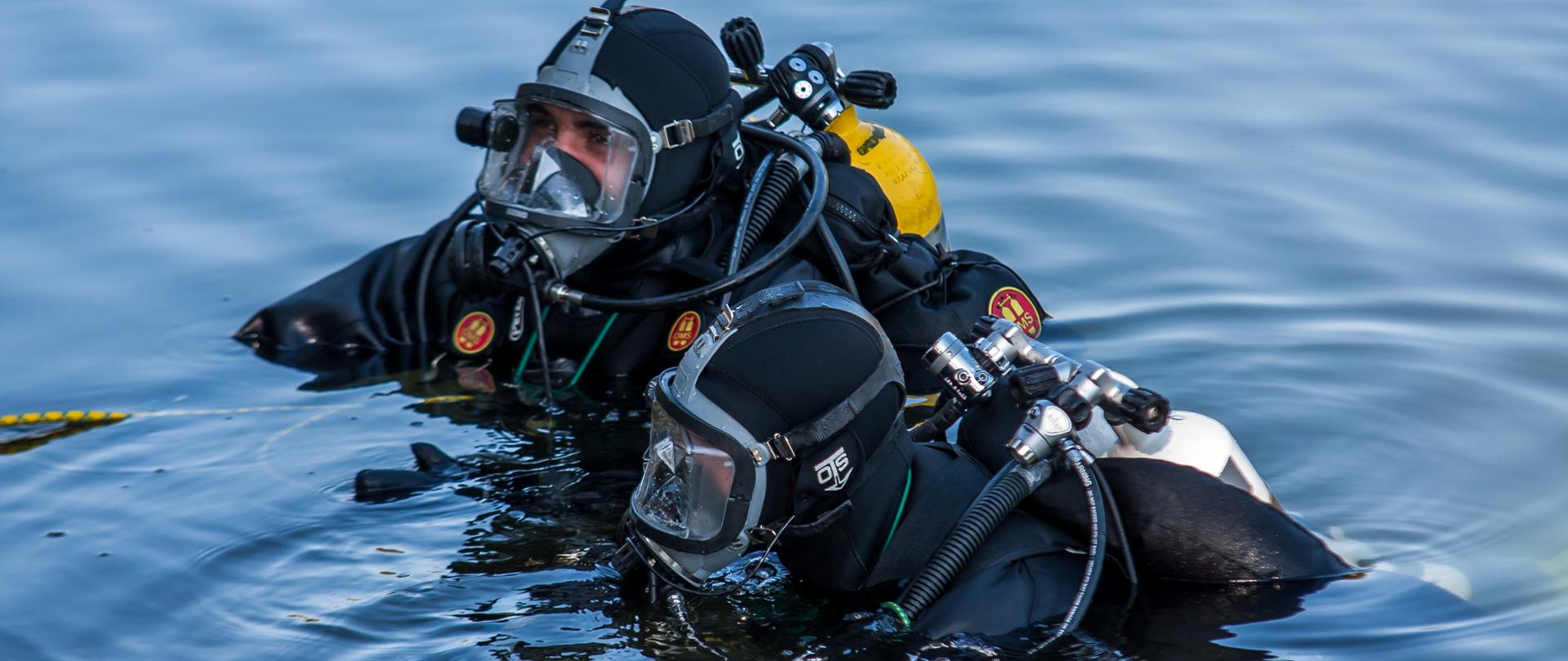 Na zdjęciu widocznych dwóch strażaków nurków Specjalistycznej Grupy Ratownictwa Wodno-Nurkowego "Opole" wykonujących działania ratownicze w wodzie. Strażacy ubrani w skafandry do pracy w wodzie z maskami pełnotwarzowymi, jacketami i butlami z powietrzem. Strażacy zanurzeni po pierś w wodzie.