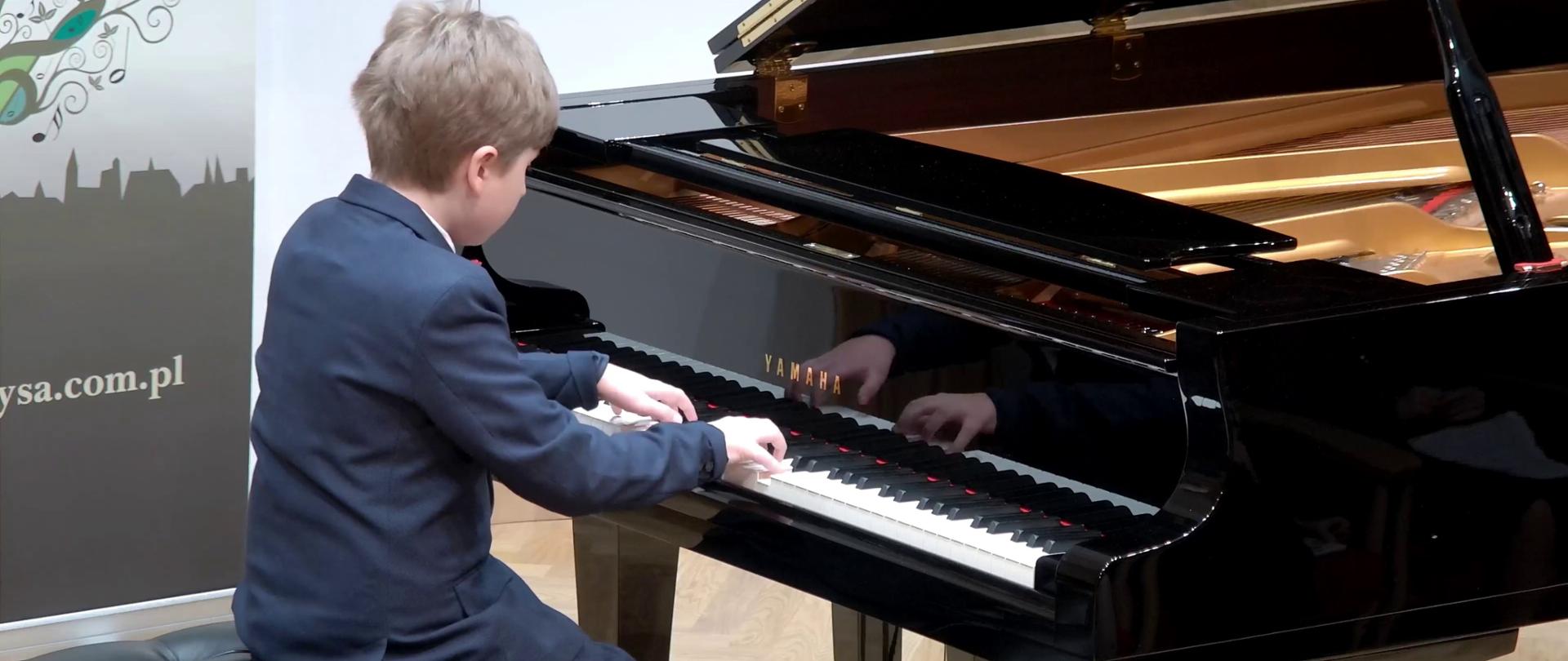 Zdjęcie przedstawia młodego pianistę grającego na dużym czarnym fortepianie. 