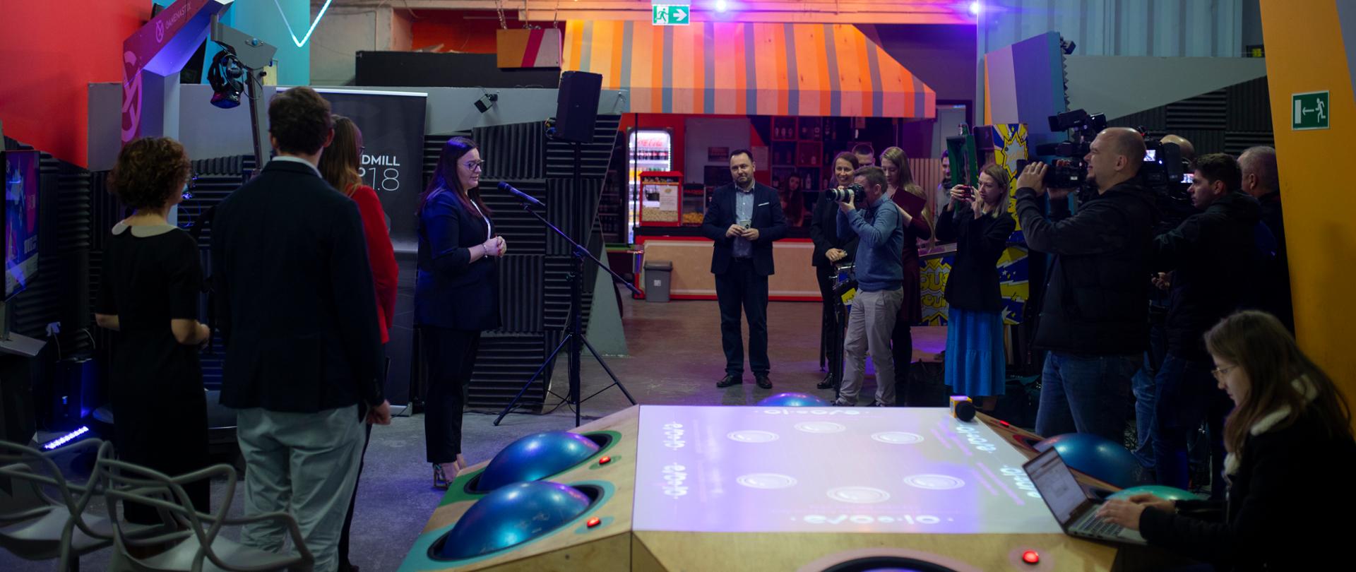 Wnętrze hali, na środku sprzęt do gier VR, po lewej stronie minister Jarosińska-Jedynak, po prawej dziennikarze z kamerami.