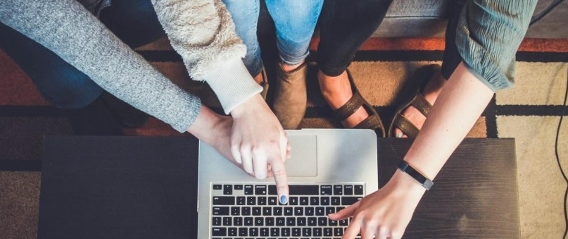 Dwie osoby współpracujące przy laptopie na kanapie; jedna wskazuje ekran, druga pisze na klawiaturze; widoczny dywan i fragment kanapy.