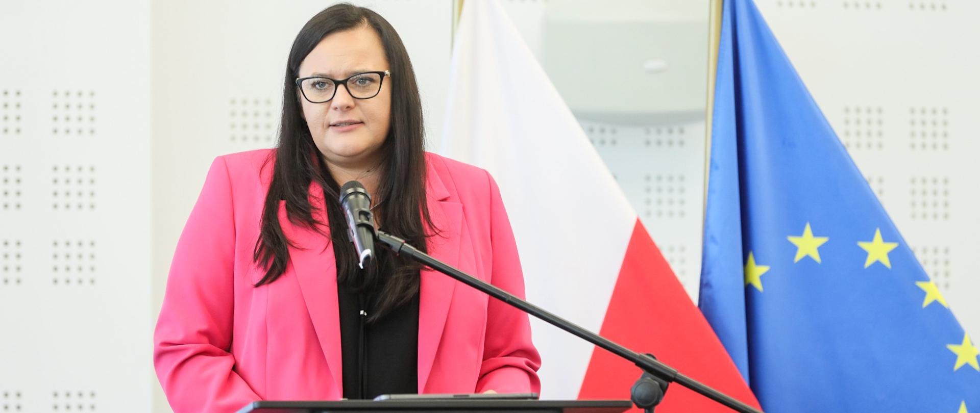 Wiceminister Małgorzata Jarosińska-Jedynak w mównicy z mikrofonem na tle flag polski i UE