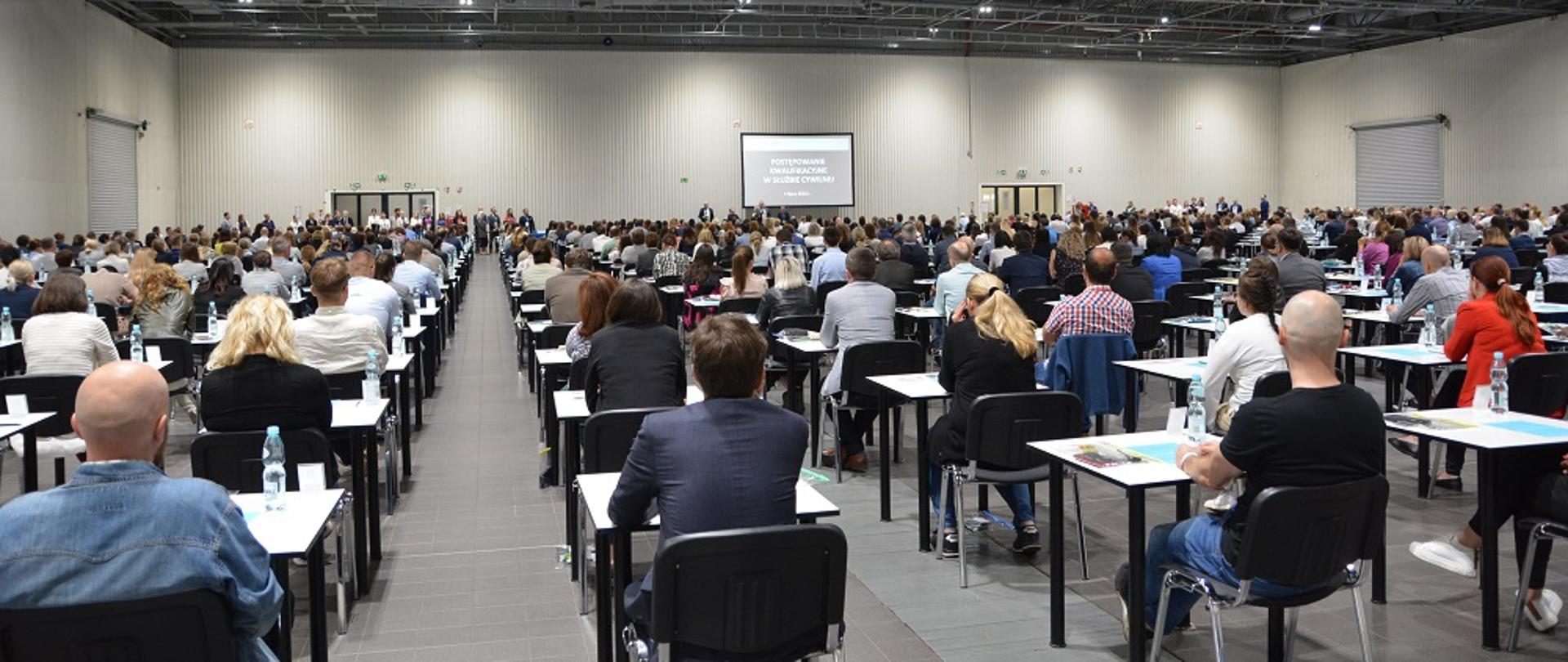 Widok sali egzaminacyjnej. Zdjęcie zza pleców uczestników. Osoby siedzą przy pojedynczych stołach odsuniętych od siebie.