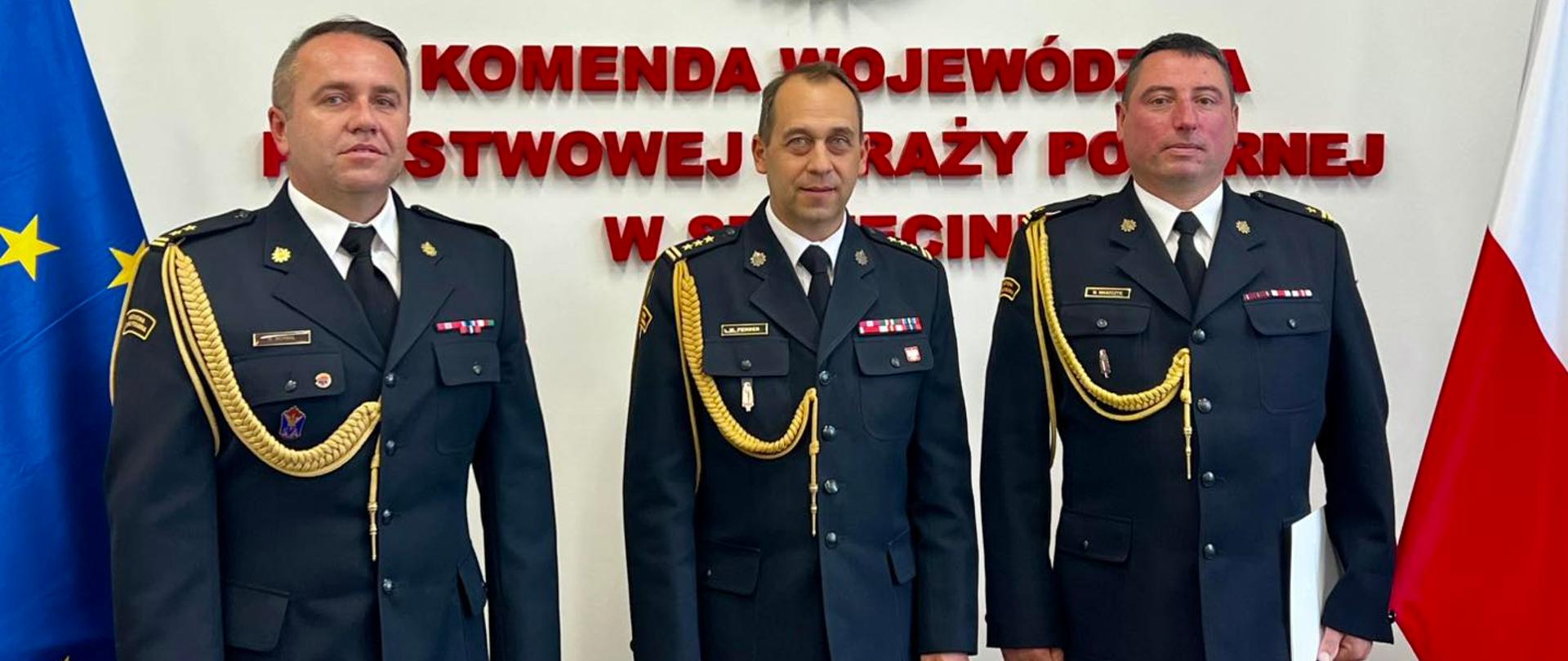 Zdjęcie przedstawia trzech funkcjonariuszy Państwowej Straży Pożarnej, stojących na tle białej ściany.
Wszyscy trzej mają ciemne mundury z jasnymi insygniami i sznurami. Na lewej piersi każdego z nich widoczne są przypięte medale i odznaczenia.
Mężczyzna po lewej stronie kadru patrzy wprost w obiektyw. Ma na sobie mundur ze złotymi sznurami.
Mężczyzna w środku również patrzy prosto na wprost. On również ma na sobie złote sznury.
Mężczyzna po prawej stronie kadru spogląda w obiektyw. On także ma na sobie złote sznury.
Za ich plecami, na ścianie, widoczny jest duży, czerwono-biały napis: "KOMENDA WOJEWÓDZKA PAŃSTWOWEJ STRAŻY POŻARNEJ W SZCZECINIE". Powyżej napisu znajduje się logo Państwowej Straży Pożarnej, przedstawiające orła w koronie na tle płomieni. Po lewej stronie zdjęcia widać fragment flagi Unii Europejskiej, a po prawej fragment flagi Polski
