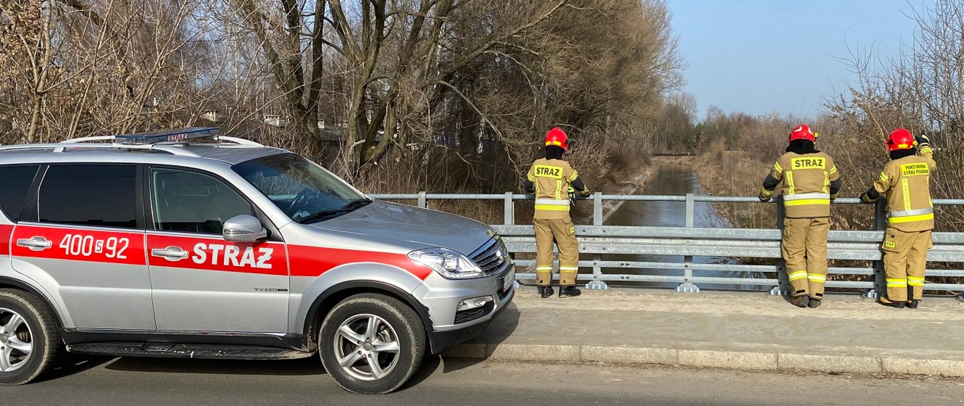 Zdjęcie przedstawia trzech strażaków w ubraniach specjalnych stojących na moście przy metalowych barierach i monitorujących poziom wody w rzece. Po lewj stronie na moście stoi samochód operacyjny Komendy Powiatowej Państwowej Straży Pożarnej