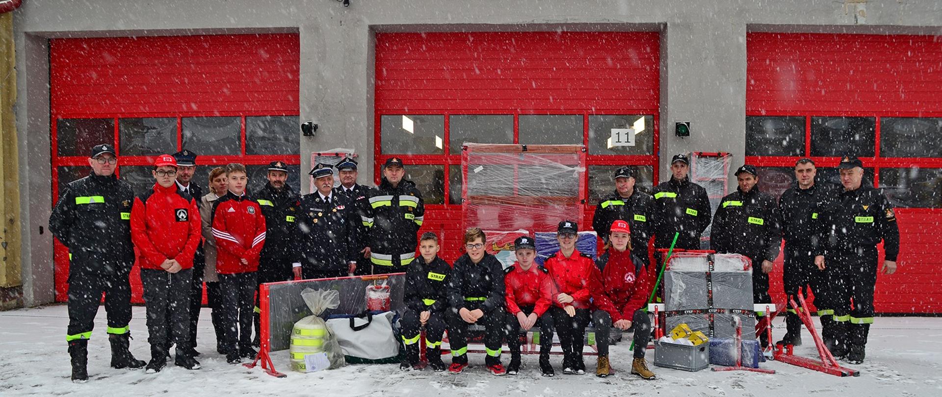 Zdjęcie grupowe. Komendant KP PSP w Chojnicach, funkcjonariusze KP PSP w Chojnicach, druhowie z OSP oraz dzieci z MDP pozują do zdjęcia z elementami torów dla Młodzieżowych Drużyn Pożarniczych, w tle czerwone bramy wjazdowe do remizy.
