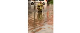 Zdjęcie przedstawia funkcjonariusza Komendy Powiatowej Państwowej Straży Pożarnej w Gołdapi, który wyposażony w długie gumowce kroczy przez jedną z posesji w gminie Gołdap, która znalazła się pod wodą. Woda sięga strażakowi do wysokości kolan. W tle widać samochód, którego tylny zderzak dotyka lustra wody