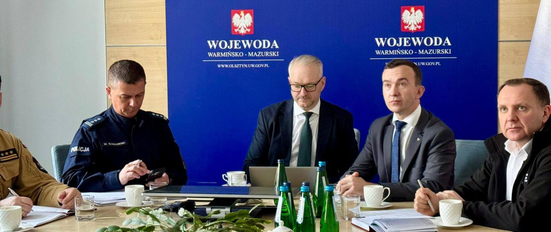 4 mężczyzn siedzi przy stole konferencyjnym na tle ścianki Wojewody Warmińsko-Mazurskiego