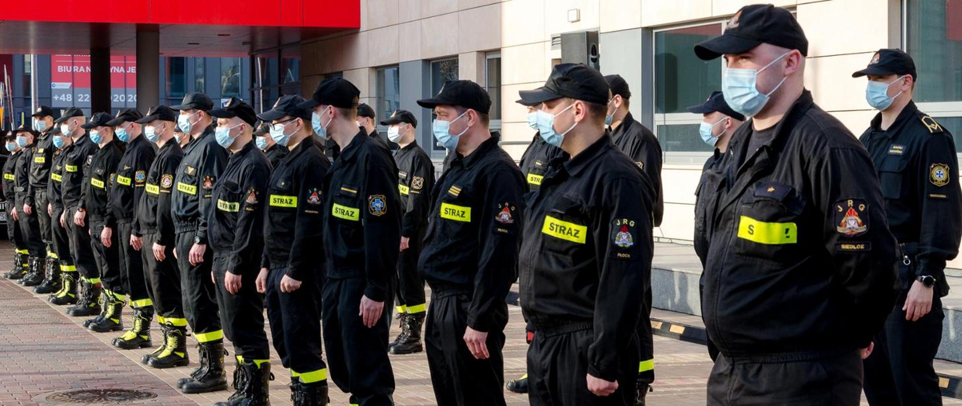 Strażacy w maseczkach ochronnych, stojący w dwuszeregu. W tle budynek KW PSP w Warszawie