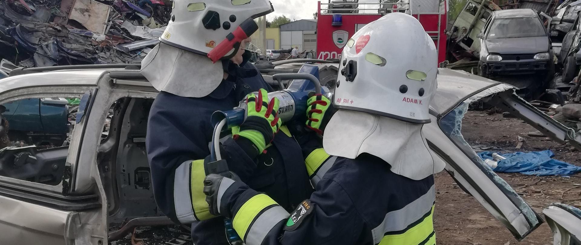 Na złomowisku dwóch strażaków przecina karoserię samochodu osobowego