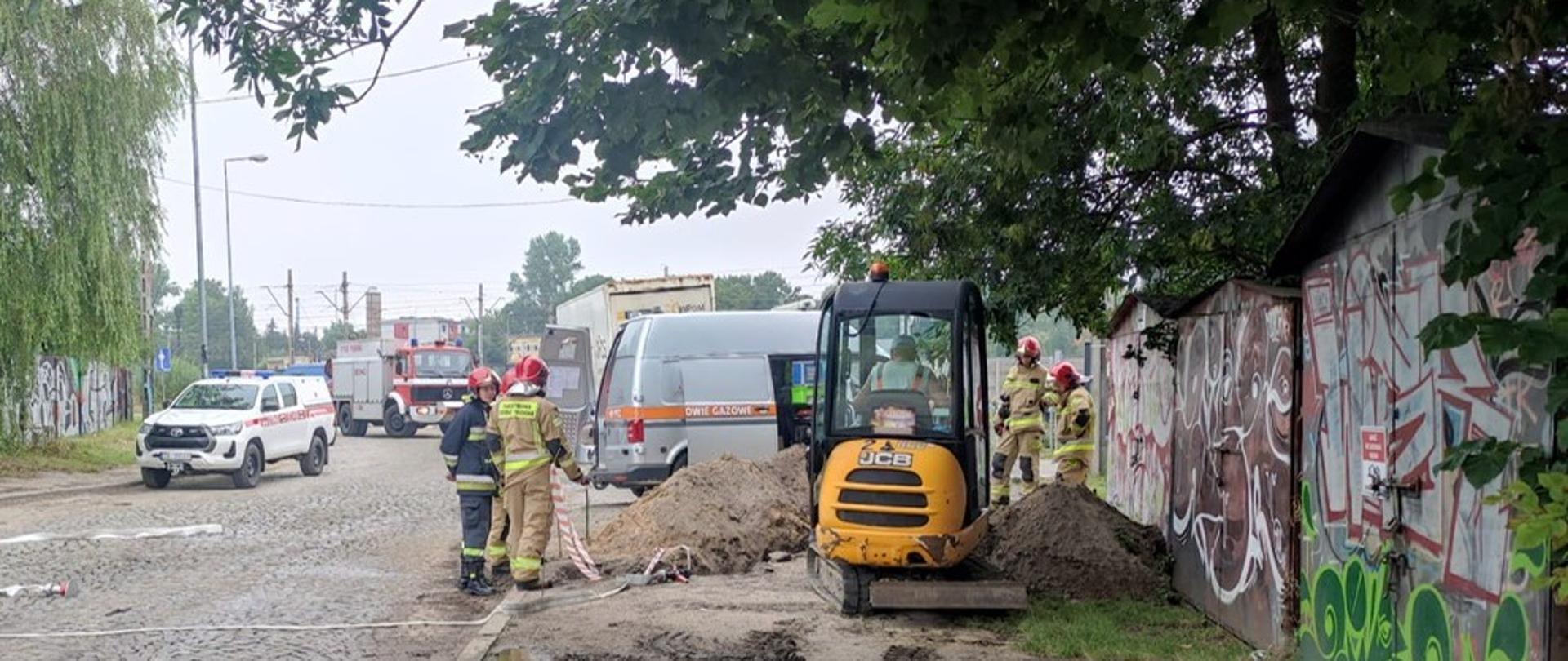 Zdjęcie przedstawia miejsce gdzie powstał wyciek gazu z uszkodzonego gazociągu. Przy pryzmie wykopanej ziemi stoi 5 strażaków, przed wykopem widać koparkę a w tle samochód Pogotowia Gazowego. W głębi ulicy stoją zaparowane 2 samochody pożarnicze