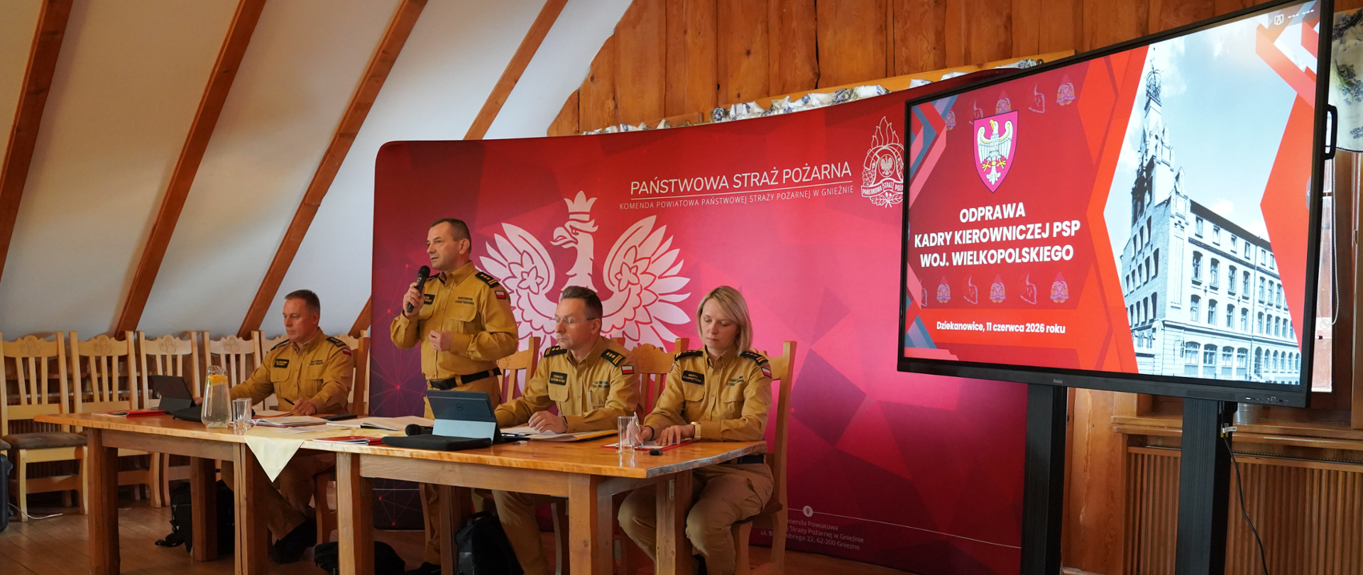 Funkcjonariusze Państwowej Straży Pożarnej podczas odprawy kadry kierowniczej PSP województwa wielkopolskiego w Dziekanowicach. Jeden ze strażaków przemawia do uczestników spotkania, siedzących przy stole konferencyjnym. W tle widoczna jest ścianka z logo PSP oraz ekran prezentacyjny z informacją o odprawie.