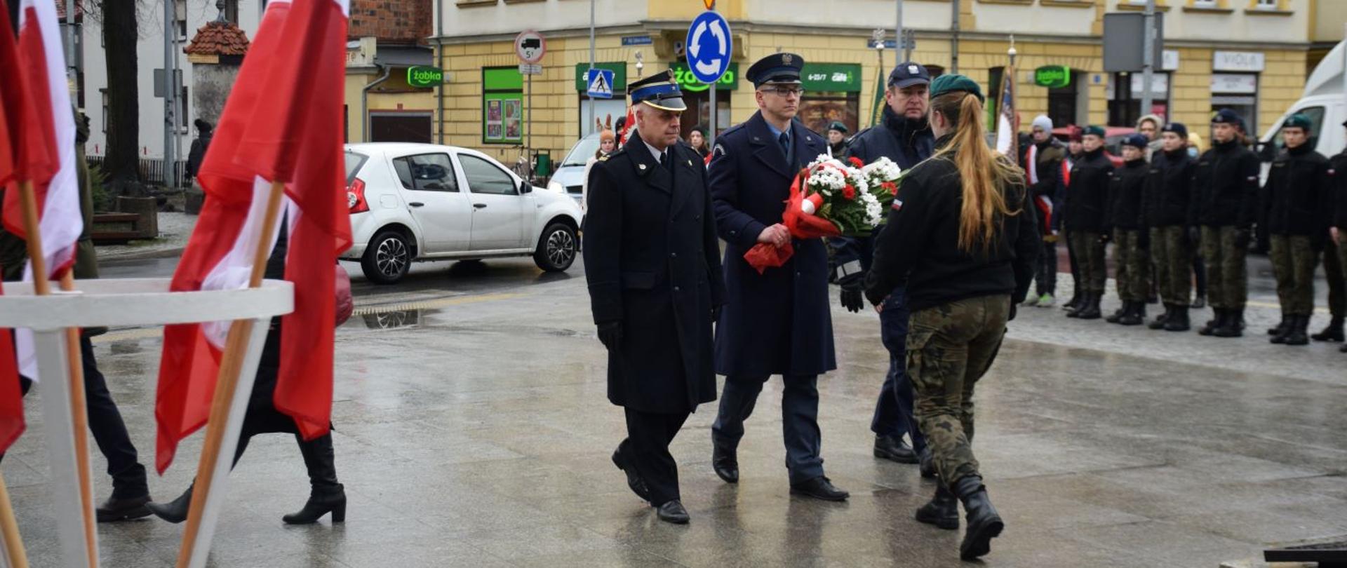 Rocznica powrotu Rawicza do Odrodzonej Polski. Plac przed pomnikiem Żołnierza Polskiego. Delegacja rawickich służb mundurowych składa wiązankę kwiatów. Z lewej strony stojak z biało-czerwonymi flagami.