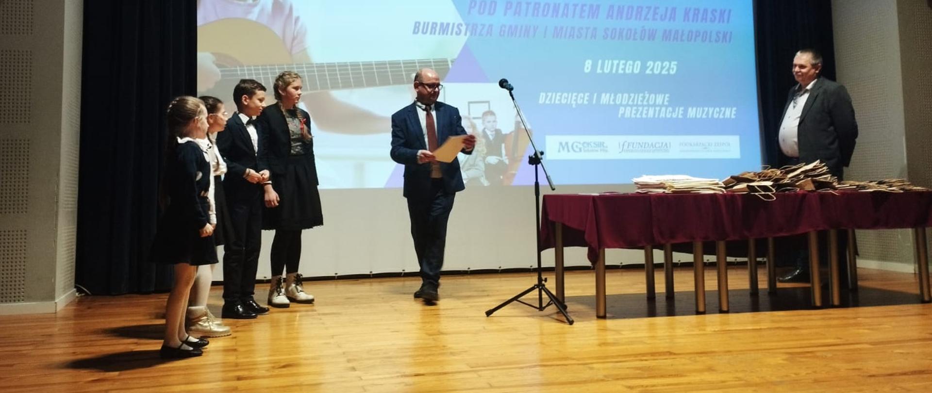 Na zdjęciu widać od lewej czworo uczniów- stojących na scenie, a obok nich mężczyznę wręczającego dyplom. Po prawej stronie stoją stoliki wraz z nagrodami oraz członek Jury.