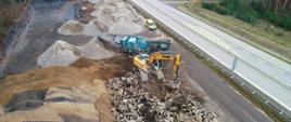 Prace rozbiórkowa na jedni południowej drogi. Pracujące maszyny, pryzmy kruszyw i połamanego betonu starej nawierzchni drogi. Jezdnia północna czynna dla ruchu z namalowanym żółtym oznakowaniem poziomym.