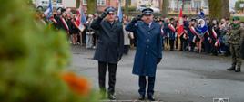 Komendant powiatowy Państwowej Straży Pożarnej oraz komendant powiatowy Policji stoją obok siebie i salutują podczas uroczystości patriotycznej w parku. Za nimi znajduje się duża grupa uczestników oraz liczne poczty sztandarowe niosące biało-czerwone i szkolne flagi. Po prawej stronie widoczny jest żołnierz w zielonym beretcie.