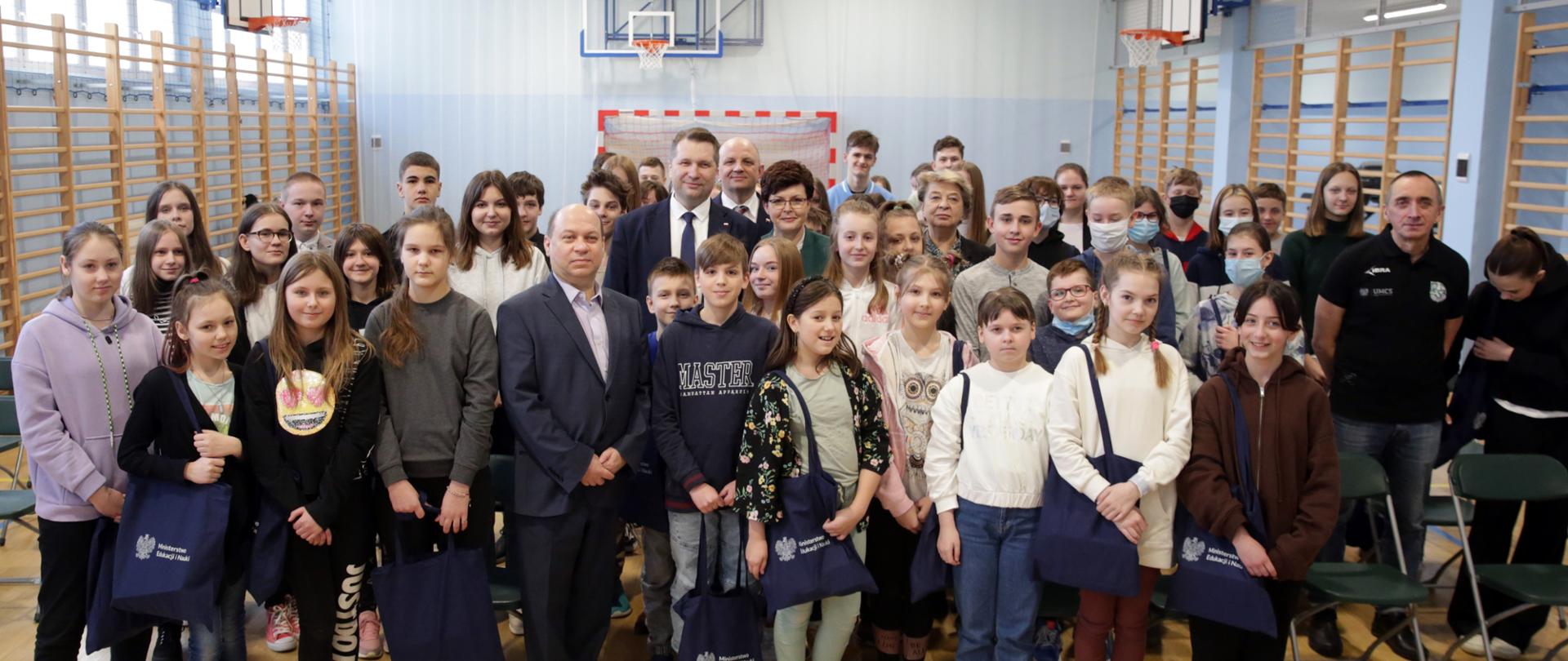 Sala gimnastyczna, grupa uczniów pozujących do tzw. family foto. Dzieci trzymają w rękach granatowe torby. W środku minister edukacji i nauki. 