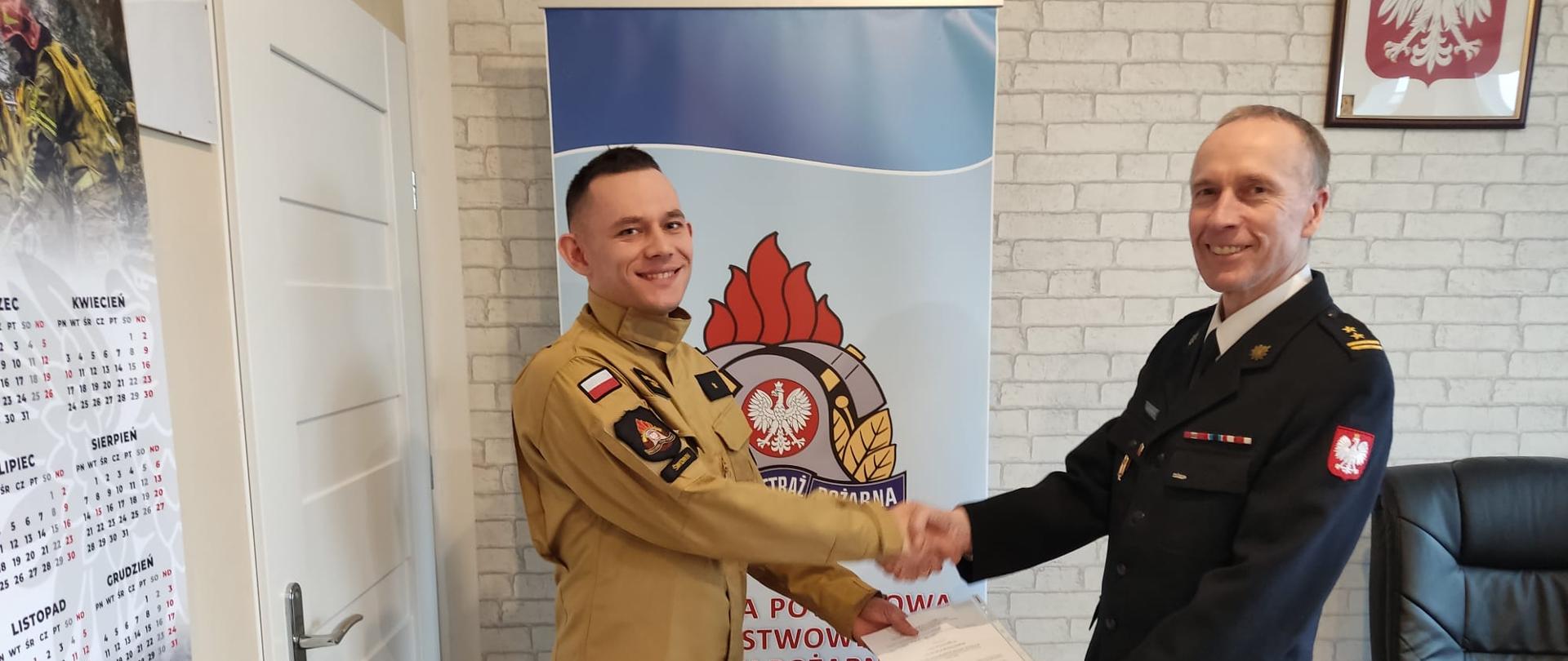 Zdjęcie przedstawia dwóch mężczyzn jeden ubrany w czarny mundur służbowe drugi w piaskowym mundur, jeden z nich trzyma w rękach dokumenty. Za nimi widnieje baner z logiem PSP oraz napisem: "Komenda Powiatowa Państwowej Straży Pożarnej w Świdwinie". Za banerem widzimy ścianę z tapetą w szare cegły. Na ścianie wisi Godło Rzeczypospolitej Polskiej. Zdjęcie wykonano w gabinecie Komendanta Powiatowego PSP w Świdwinie.