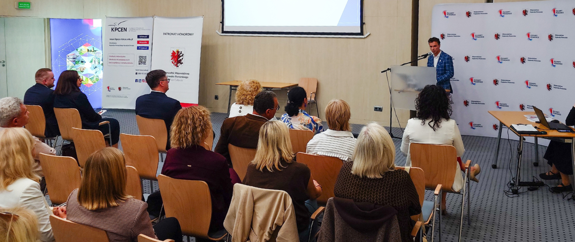 Rafał Kochanowski z Wojewódzkiej Stacji Sanitarno-Epidemiologicznej w Bydgoszczy wygłasza prelekcję podczas konferencji „Zdrowie na co dzień” w Urzędzie Marszałkowskim w Toruniu. Stoi za mównicą z mikrofonem, zwrócony w stronę uczestników siedzących w sali konferencyjnej. W tle widoczne są banery organizatorów i patronów wydarzenia.
