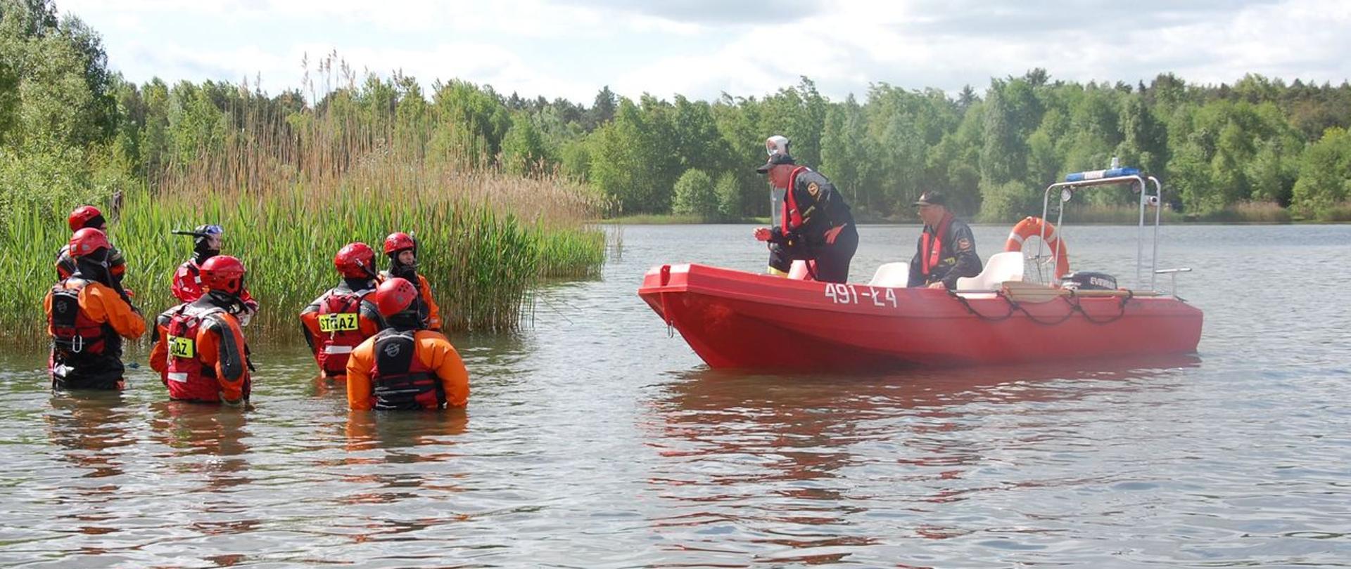 Zdjęcie przedstawia uczestników szkolenia w trakcie instruktarzu odnośnie sposobów prowadzenia działań na akwenach wodnych. Strażacy stoją po pas w wodzie. Ćwiczący ubrani są w suche skafandry ratownicze oraz kamizelki asekuracyjne. Głowy zabezpieczone mają kaskami ochronnymi z przeznaczeniem do ratownictwa wodnego. Dowódca Specjalistycznej Grupy Wodno-Nurkowej prowadzący zajęcia przebywa na łodzi wraz ze sternikiem łodzi. Obsada łodzi ratowniczej zabezpieczona jest manualnymi kamizelkami ratowniczymi.
