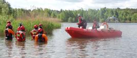 Zdjęcie przedstawia uczestników szkolenia w trakcie instruktarzu odnośnie sposobów prowadzenia działań na akwenach wodnych. Strażacy stoją po pas w wodzie. Ćwiczący ubrani są w suche skafandry ratownicze oraz kamizelki asekuracyjne. Głowy zabezpieczone mają kaskami ochronnymi z przeznaczeniem do ratownictwa wodnego. Dowódca Specjalistycznej Grupy Wodno-Nurkowej prowadzący zajęcia przebywa na łodzi wraz ze sternikiem łodzi. Obsada łodzi ratowniczej zabezpieczona jest manualnymi kamizelkami ratowniczymi.
