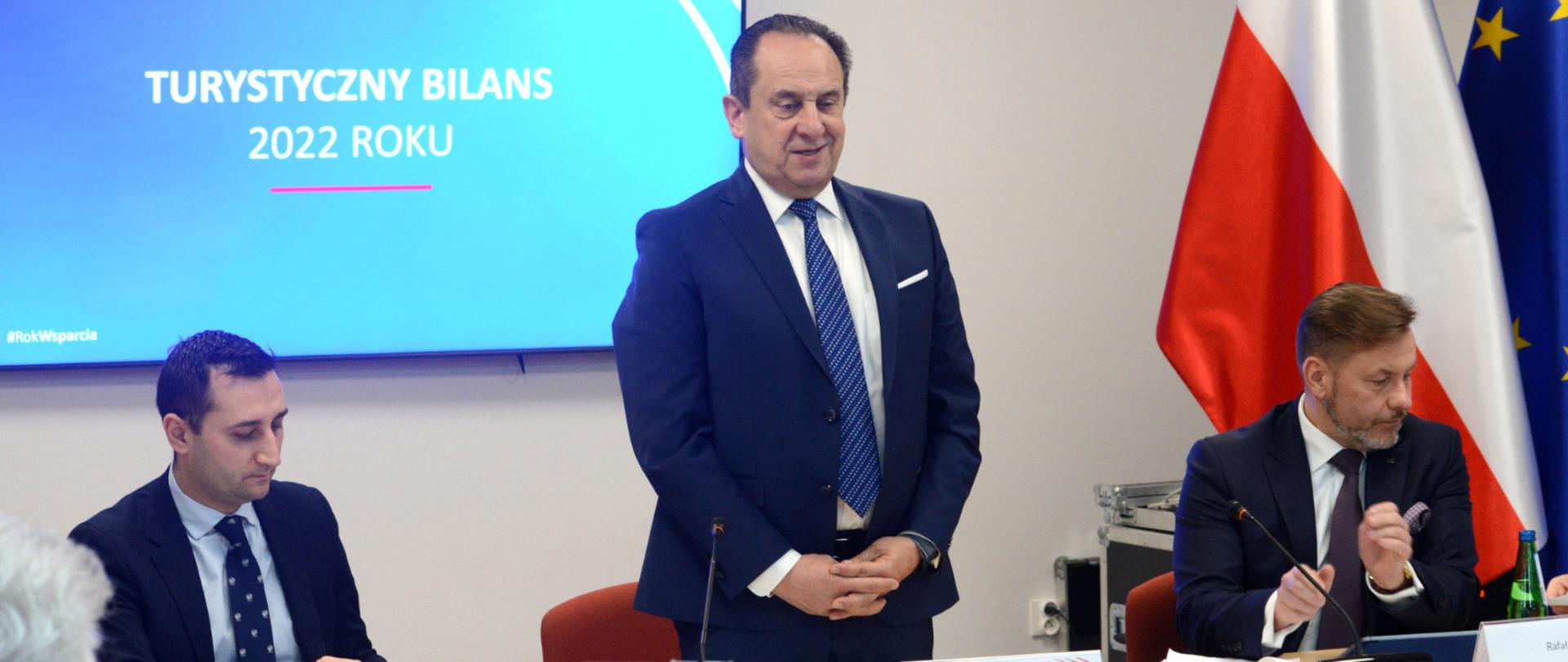 Na zdjęciu znajduje się trzech elegancko ubranych mężczyzn. Jeden z nich stoi i jest w trakcie wystąpienia publicznego. W tle znajdują się flagi: Polski i Unii Europejskiej. Na ekranie zawieszonym na ścianie, która jest tłem zdjęcia znajduje się napis "Turystyczny Bilans 2022 roku".