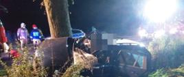 zdjęcie robione od strony pobocza na drogę. Przechylone auto oparte spodem o stojące przy drodze drzewo. Konstrukcja pojazdu widocznie pokrzywdzona. W tle stoi dwóch strażaków oraz pojazdy pożarnicze i radiowóz.