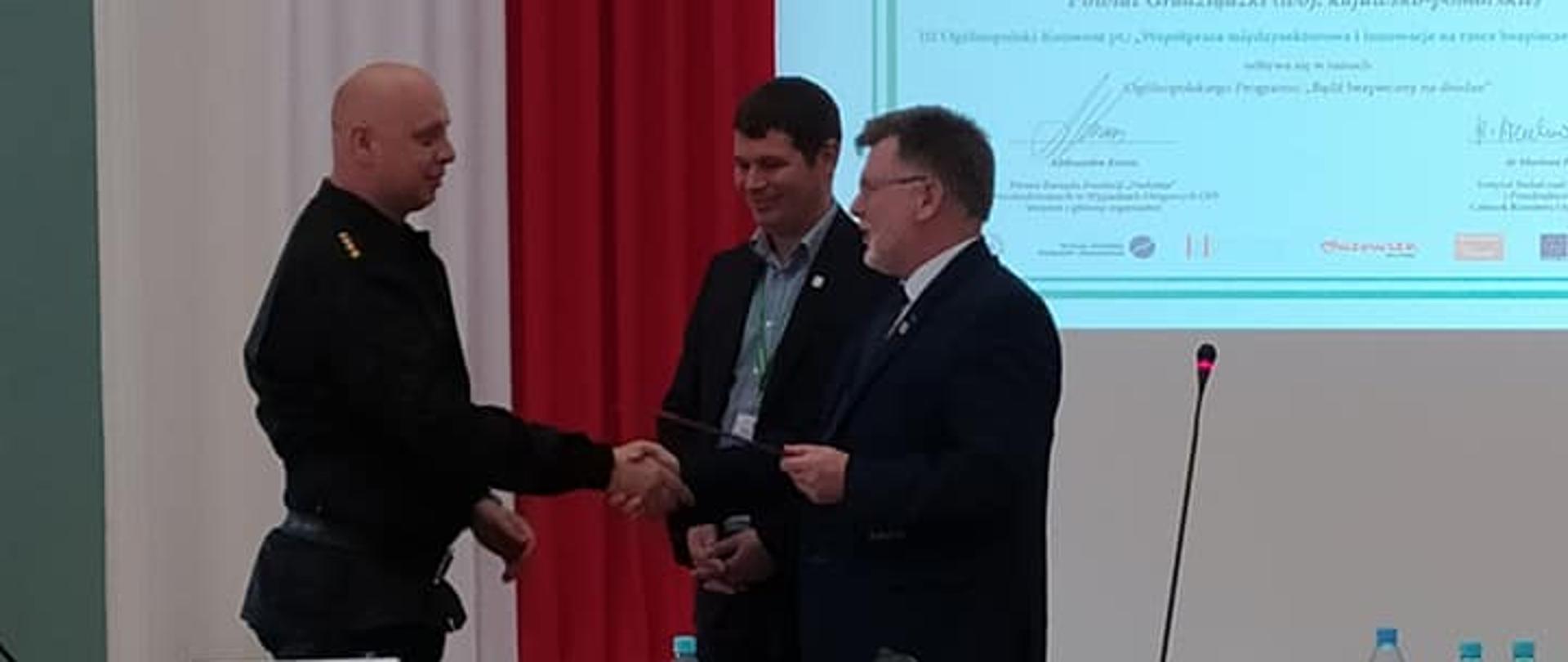 Zdjęcie przedstawia salę konferencyjną, w której za stołem stoją trzej mężczyźni. Dwaj z nich ściskają sobie dłonie. Na ścianie za nimi wyświetlana jest prezentacja multimedialna.