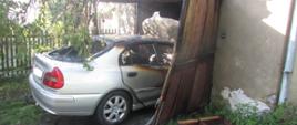 Srebrny samochód, spalony w połowie, stoi częściowo na podjeździe, a od maski w murowanym garażu z drewnianą bramą. W tle płot posesji oraz drzewa.