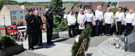 Strażacy w galowych mundurach oraz kapłan stoją przy pomniku upamiętniającym wizytę Jana Pawła II. Obok nich ustawiona jest grupa kobiet w białych bluzkach, a u stóp monumentu znajdują się wieńce i znicze. W tle widoczne są domy, wóz strażacki i zgromadzeni mieszkańcy.