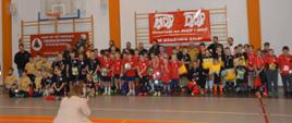 Na hali sportowej, na tle drabinek, na których umieszczone są czerwone banery z białymi napisami i logami, do wspólnego zdjęcia pozują zawodnicy, opiekunowie, ortganizatorzy i goście turnieju. Zawodnicy maja na szyjach zawieszone medale, w rękach trzymają dyplomy i nagrody. 