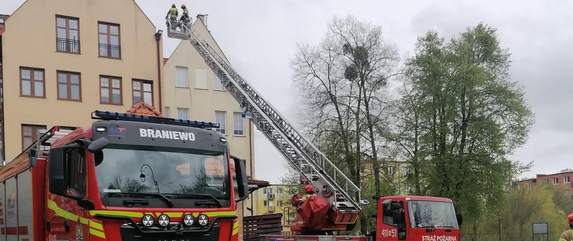 Na zdjęciu Na tle budynków z lewej strony stoi czerwony samochód strażacki i obok drabina mechaniczne strażacka. W koszu drabiny strażacy.