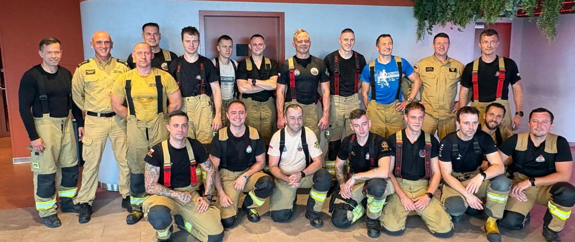 Grupa dziewiętnastu strażaków w piaskowo-brązowych oraz czarnych mundurach bojowych pozuje do wspólnego zdjęcia w nowoczesnym wnętrzu.