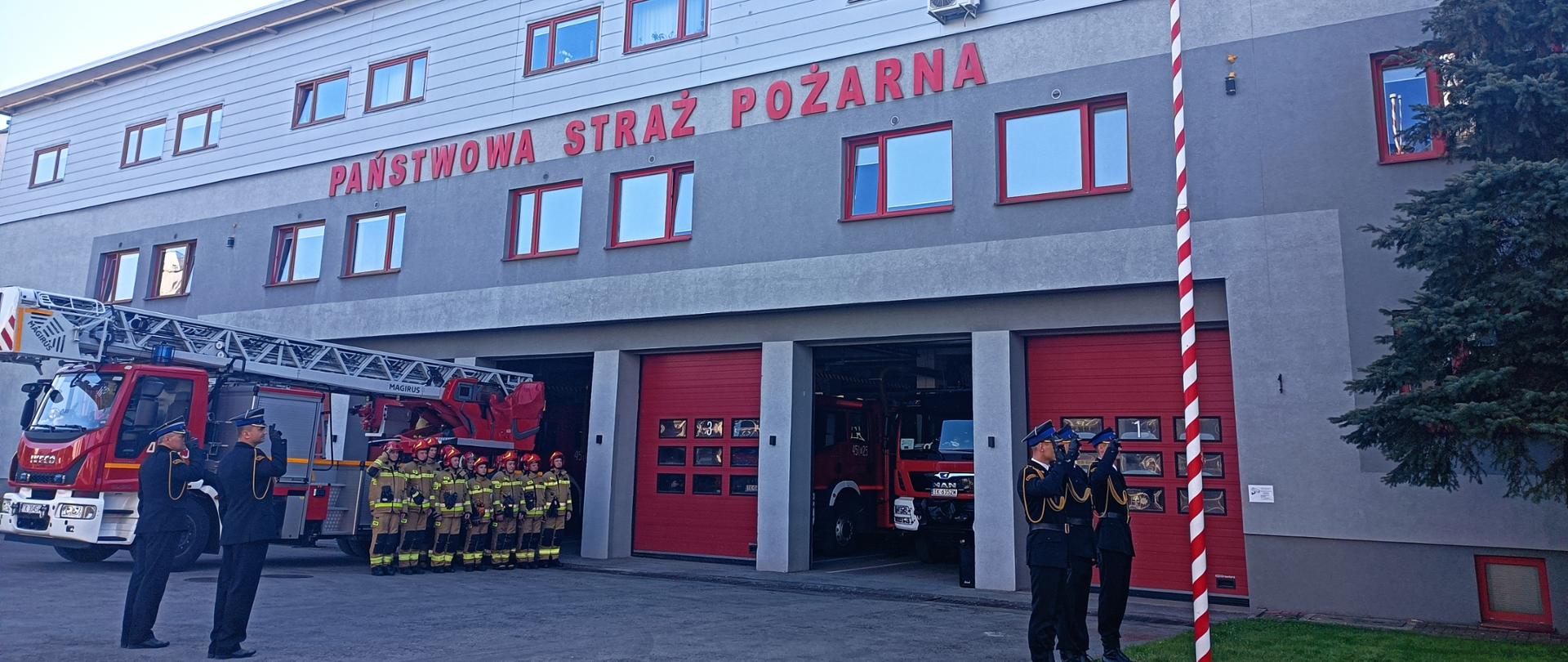 Zdjęcie wykonane przed siedzibą KP PSP w Ostrowcu Św. podczas uroczystej zbiórki. Na zdjęciu po lewej stronie na baczność stoi kadra dowódcza KP PSP w Ostrowcu Św. Za strażakami stojącymi w szeregu zaparkowany jest samochód pożarniczy drabina. Na pierwszym planie widać salutujących strażaków przed masztem flagowym. Na maszcie flagowym zawisła właśnie flaga Polski.