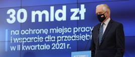 Wicepremier, minister rozwoju, pracy i technologii Jarosław Gowin stojący przy mównicy z mikrofonem, za jego placami ekran z wyświetloną informacją o pomocy 30 mld zł na ochronę miejsc pracy i wsparcie dla przedsiębiorców w II kwartale 2021 r.