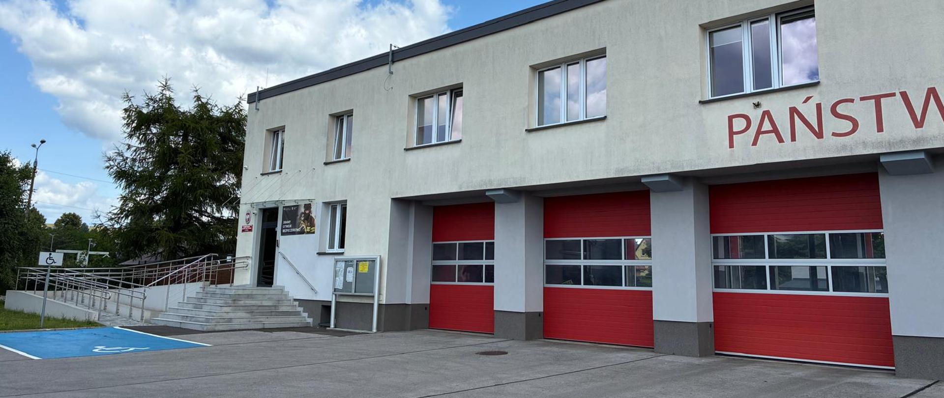 Na zdjęciu widoczny jest nowoczesny, dwukondygnacyjny budynek Komendy Miejskiej Państwowej Straży Pożarnej w Jaworznie. Po lewej stronie budynku znajduje się wejście główne z kilkoma stopniami oraz podjazdem dla osób z niepełnosprawnościami. Przed budynkiem znajduje się betonowy plac manewrowy oraz miejsce parkingowe dla osób niepełnosprawnych. Na niebie widać jasne chmury na tle błękitnego nieba.